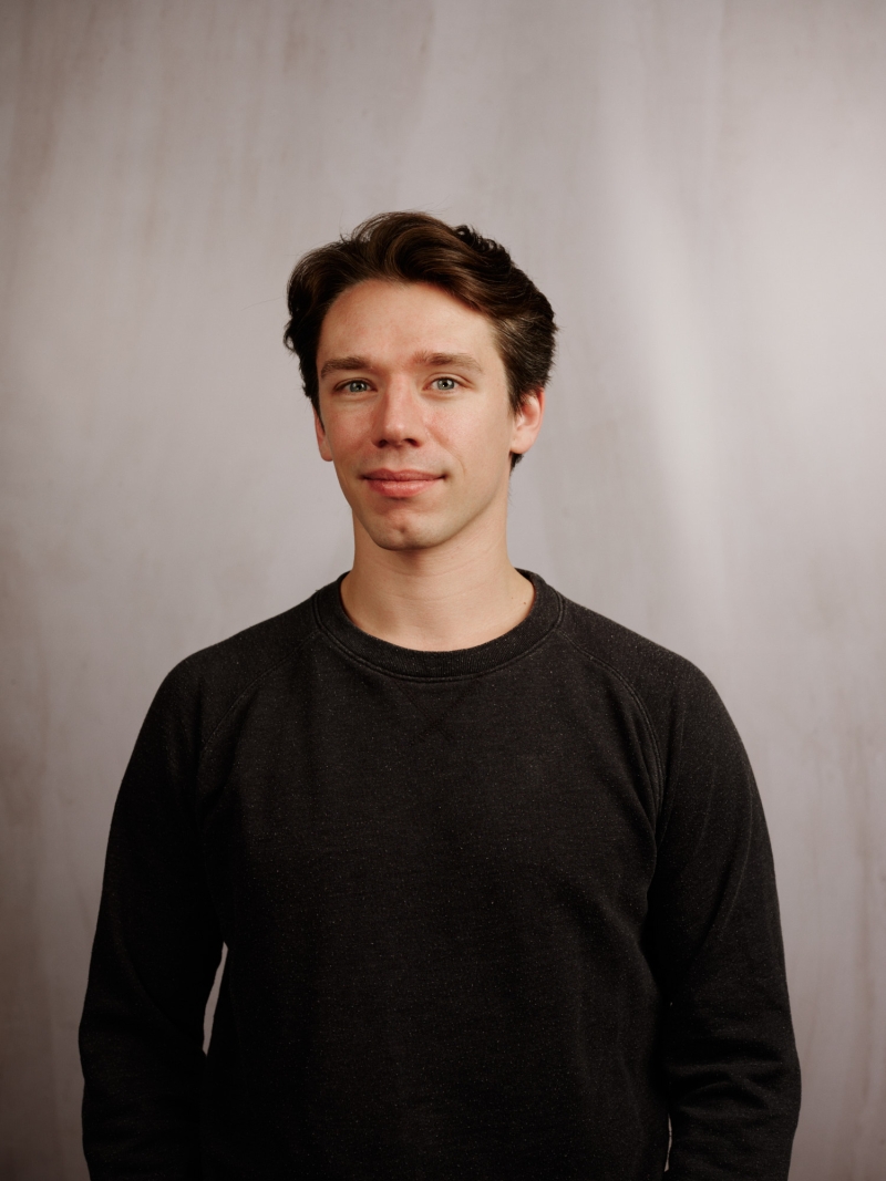 Junger Mann mit kurzen, dunklen Haaren in einem schwarzen Pullover, vor einem hellen, einfarbigen Hintergrund.