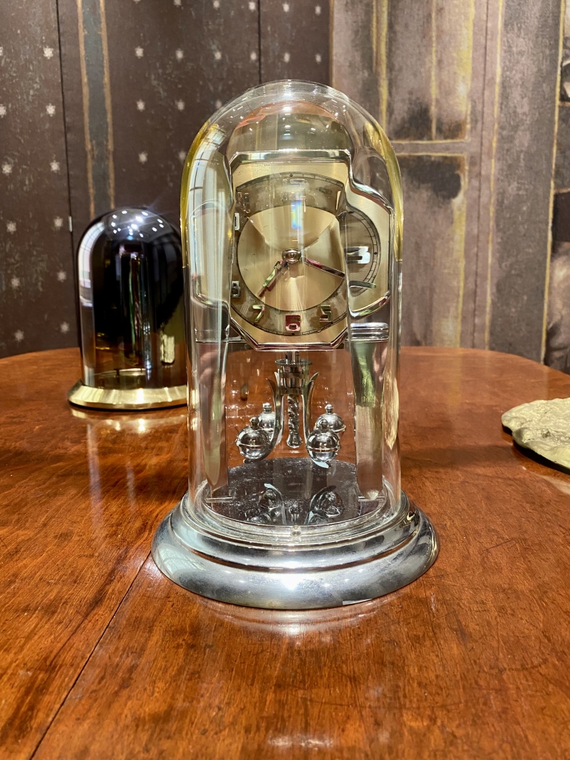 A vintage clock under a glass dome, featuring a gold face and silver base, on a wooden table with a dark background.