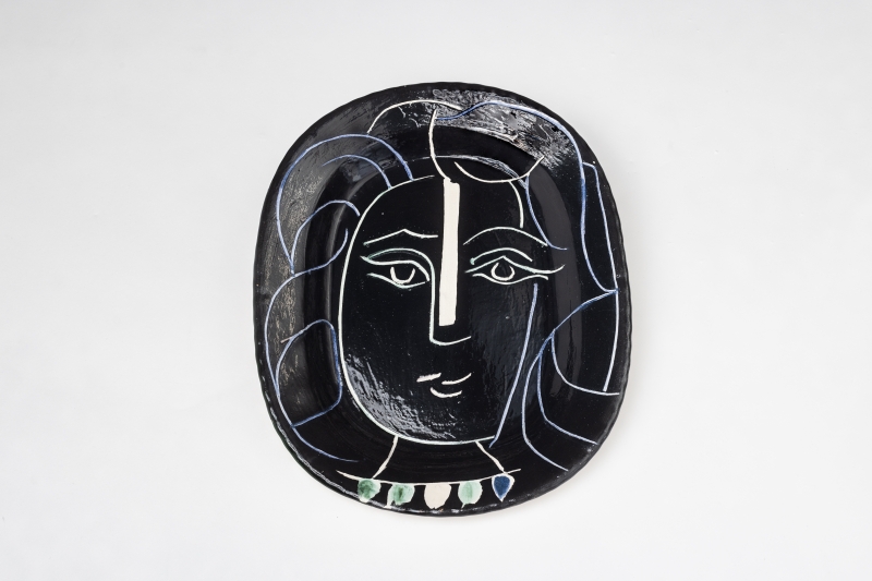 Black ceramic plate featuring a stylized face with wavy hair, outlined in white, and colorful accents at the bottom.