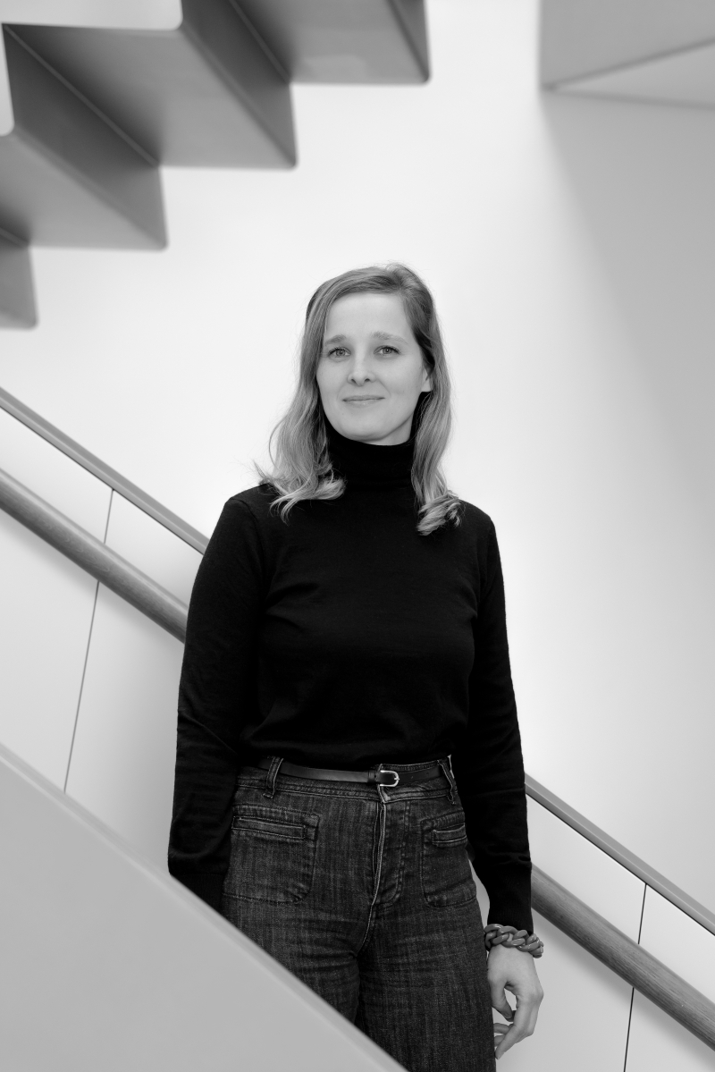 Auf dem Bild sieht man Véronique Abpurg.
Sie steht vor einer modernen Treppe mit klaren Linien.
Véronique trägt einen schwarzen Rollkragenpullover und eine dunkle Jeans.
Sie hat schulterlanges, leicht gewelltes Haar und blickt freundlich und ruhig in die Kamera.
Das Foto ist in Schwarz-Weiß aufgenommen.