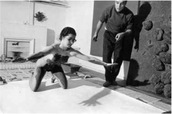 Eine nackte Frau mit Brille streckt ihren Arm auf ein großes Blatt Papier, während ein Mann daneben steht.