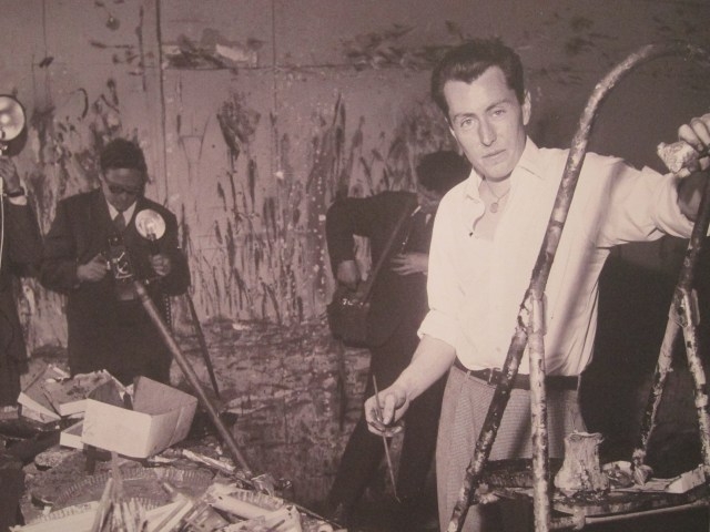 Bernard Buffet stands at an easel, surrounded by journalists, in a cluttered artist's studio with paint splatters on the walls.