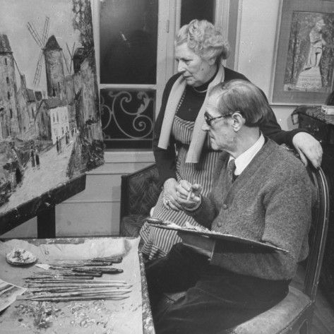 A man and woman observe a painting of a landscape with windmills, surrounded by art supplies and studio decor.