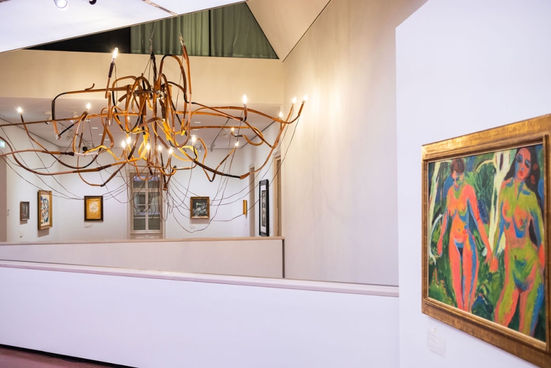 A modern chandelier with twisted branches hangs in an art gallery, featuring colorful paintings on the walls.