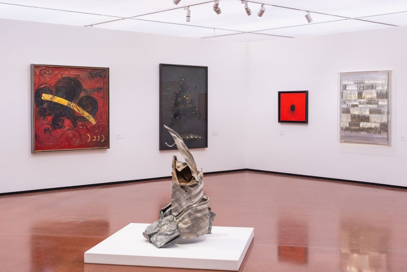 Sculpture in foreground with abstract paintings on white walls, featuring bold colors and varied textures.