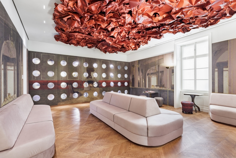 The photo shows the Tea Room in the Heidi Horten Collection museum in Vienna.
A large, red work of art by Hans Kupelwieser hangs from the ceiling.
It looks like crumpled paper or shiny fabric, but
is made of aluminum.
There are lots of round windows on the back wall - they look like portholes.
Behind them are small, precious objects from Heidi Horten's collection.
This showcase wall was designed by Markus Schinwald.
The pink seating in the middle of the room is also part of his concept.
They invite you to sit down, look and relax.
The walls are covered with large fabric pictures,
reminiscent of an old café or castle room.