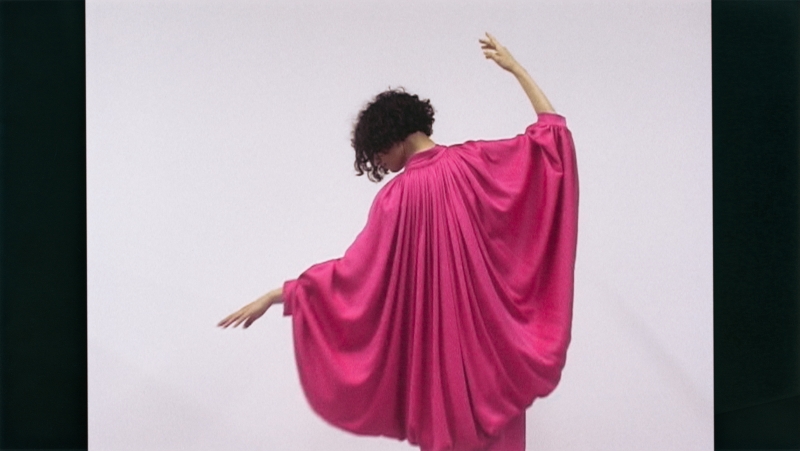 Frau in einem fließenden, pinken Kleid mit weiten Ärmeln, die sich elegant zur Seite dreht.