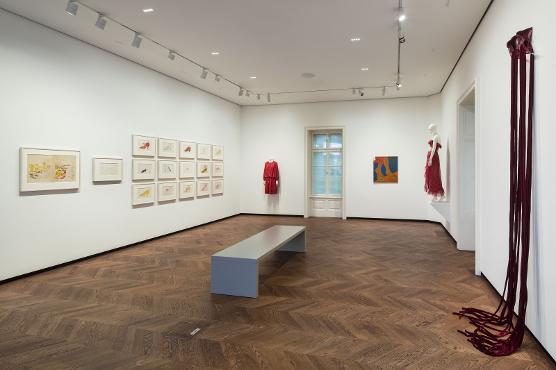 Ausstellungsraum mit Holzfußboden, Kunstwerken an Wänden, einer Bank in der Mitte und roten Kleidungsstücken.