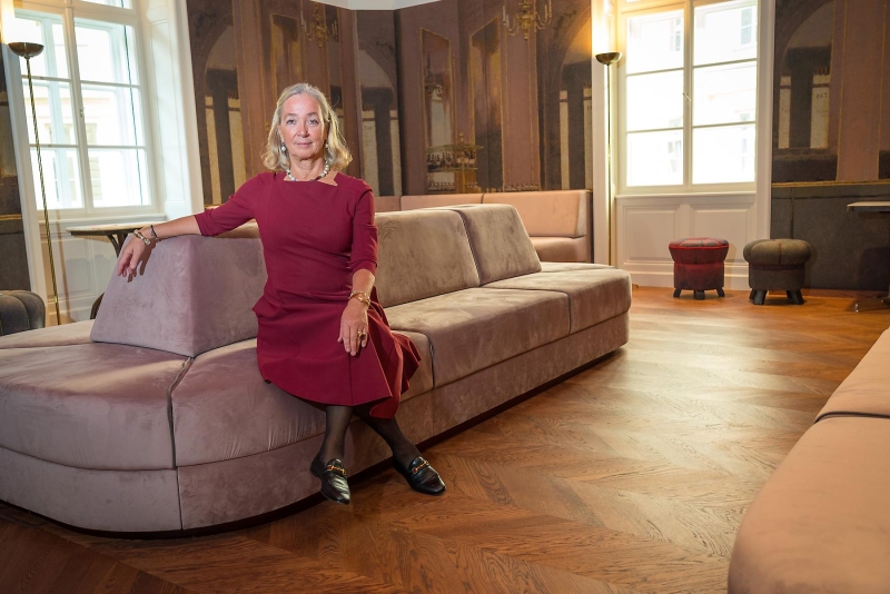 Agnes Husslein-Arco in rotem Kleid sitzt entspannt auf einer modernen, hellen Couch in einem eleganten Raum mit Holzfußboden.