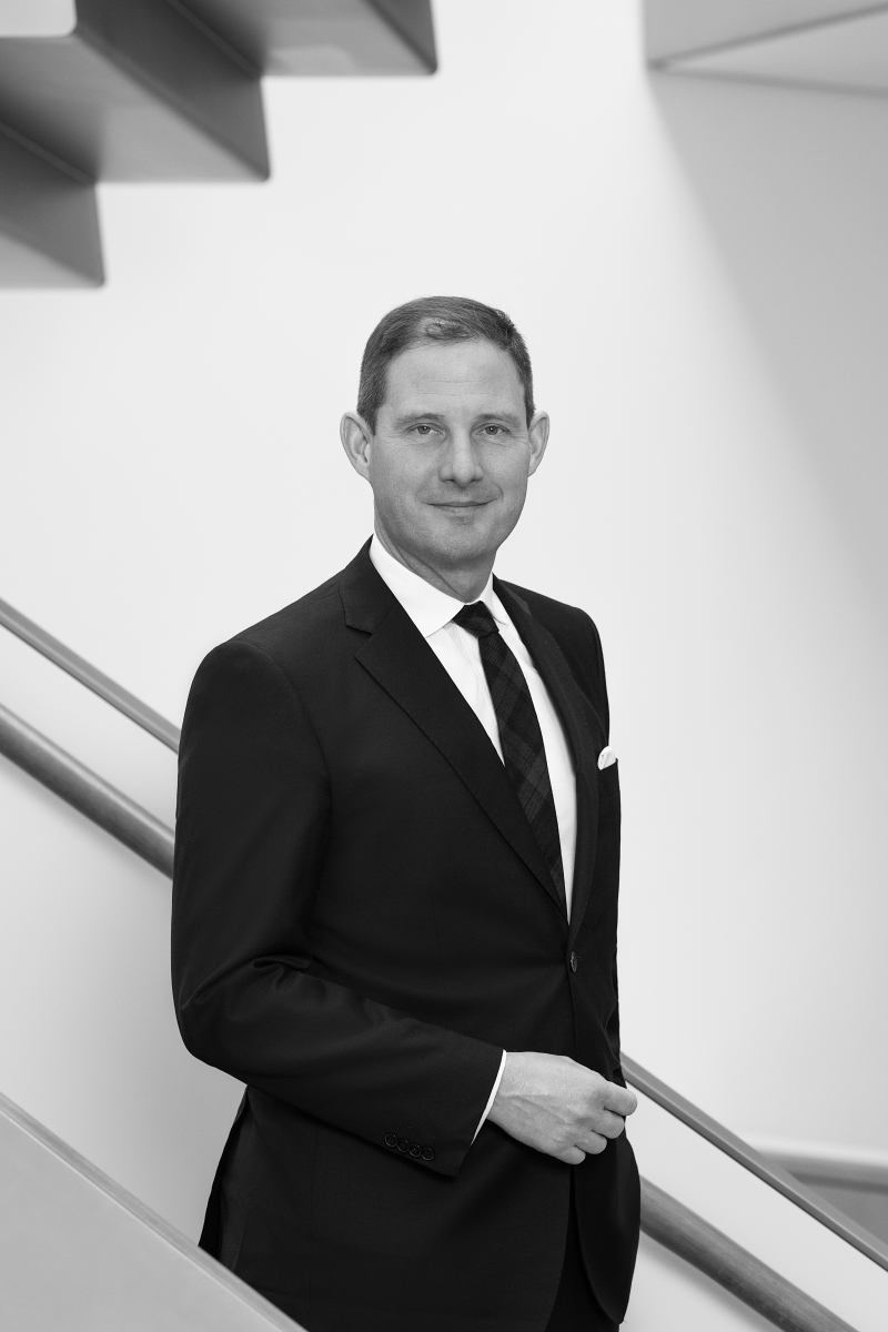 Auf dem Bild sieht man einen Mann, namens Heinz Holzinger, im Anzug.
Er trägt ein dunkles Sakko, ein weißes Hemd und eine Krawatte.
In seiner Brusttasche steckt ein weißes Tuch.
Der Mann steht vor einer modernen Treppe mit klaren Linien.
Er schaut freundlich und selbstbewusst in die Kamera.
Seine Haltung ist aufrecht und ruhig.
Das Foto ist in Schwarz-Weiß aufgenommen.