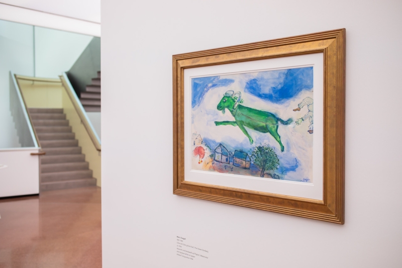 Grüne fliegende Kuh vor blauem Himmel, umgeben von Häusern und Bäumen, eingerahmt in goldenem Rahmen.