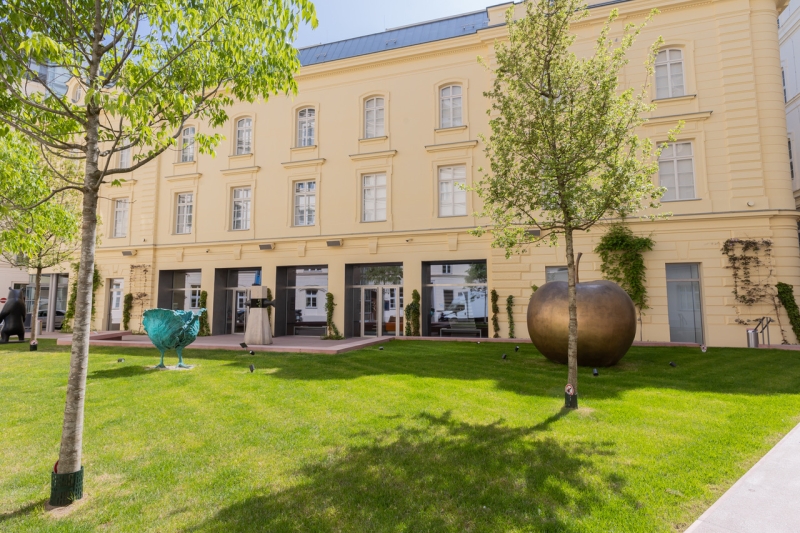 Gelbes Gebäude mit großen Fenstern, umgeben von Rasen, Bäumen und modernen Skulpturen.