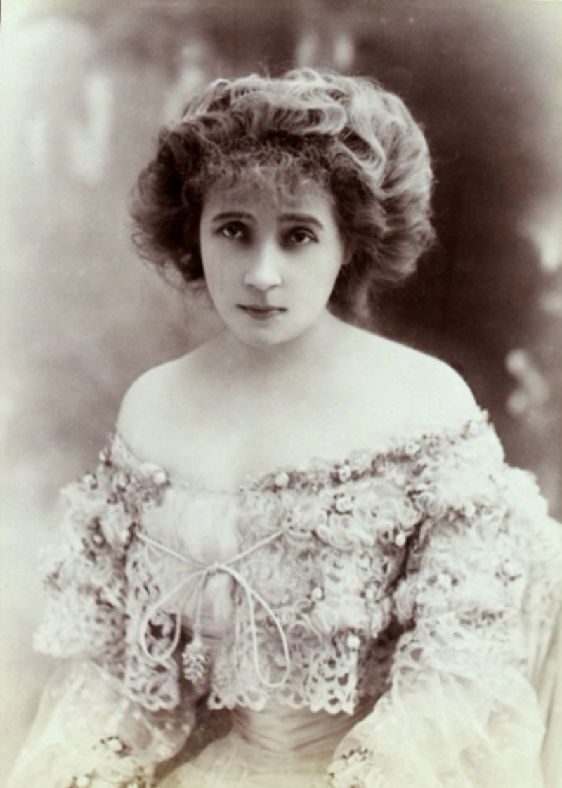 Portrait of a woman with curly hair, wearing an ornate lace dress, gazing directly at the viewer with a serious expression.