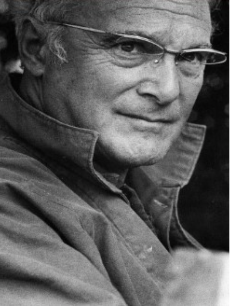 A close-up of a man with glasses, gray hair, and a thoughtful expression, wearing a collared jacket.