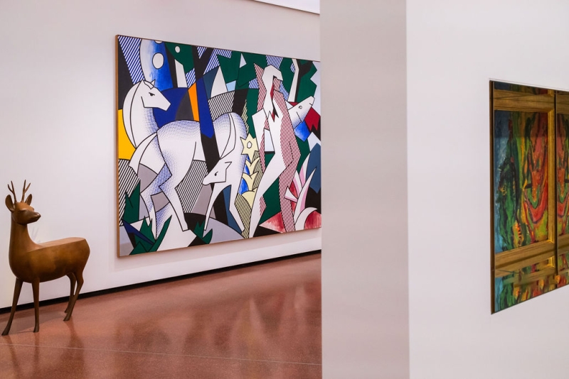 A bronze deer sculpture stands beside a colorful abstract painting of horses and foliage in a gallery setting.