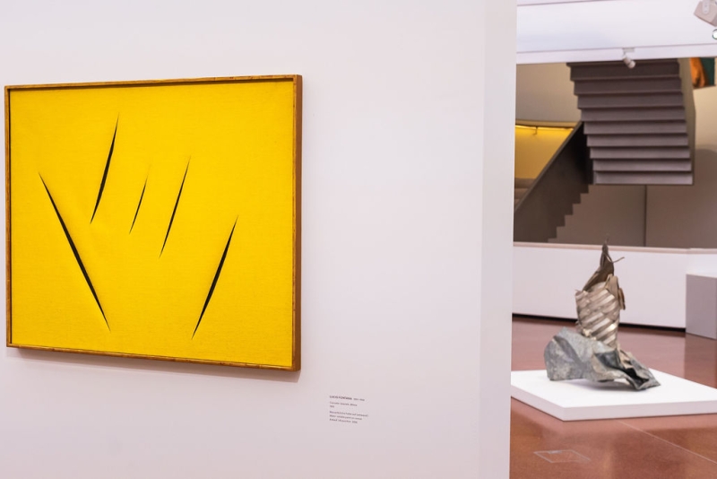 Abstract yellow artwork with black slashes, displayed in a gallery with a metallic sculpture in the background.