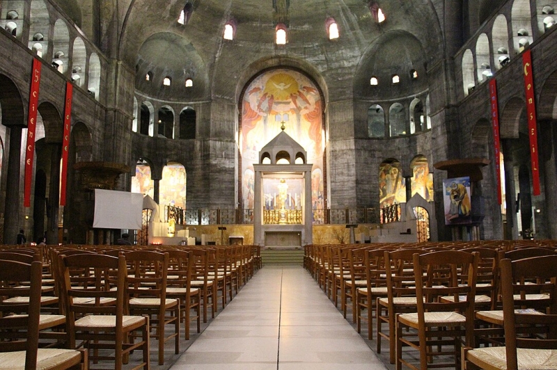 Innenansicht der Église du Saint-Esprit mit Holzstühlen, bunten Wandmalereien und einem Altar im Hintergrund.