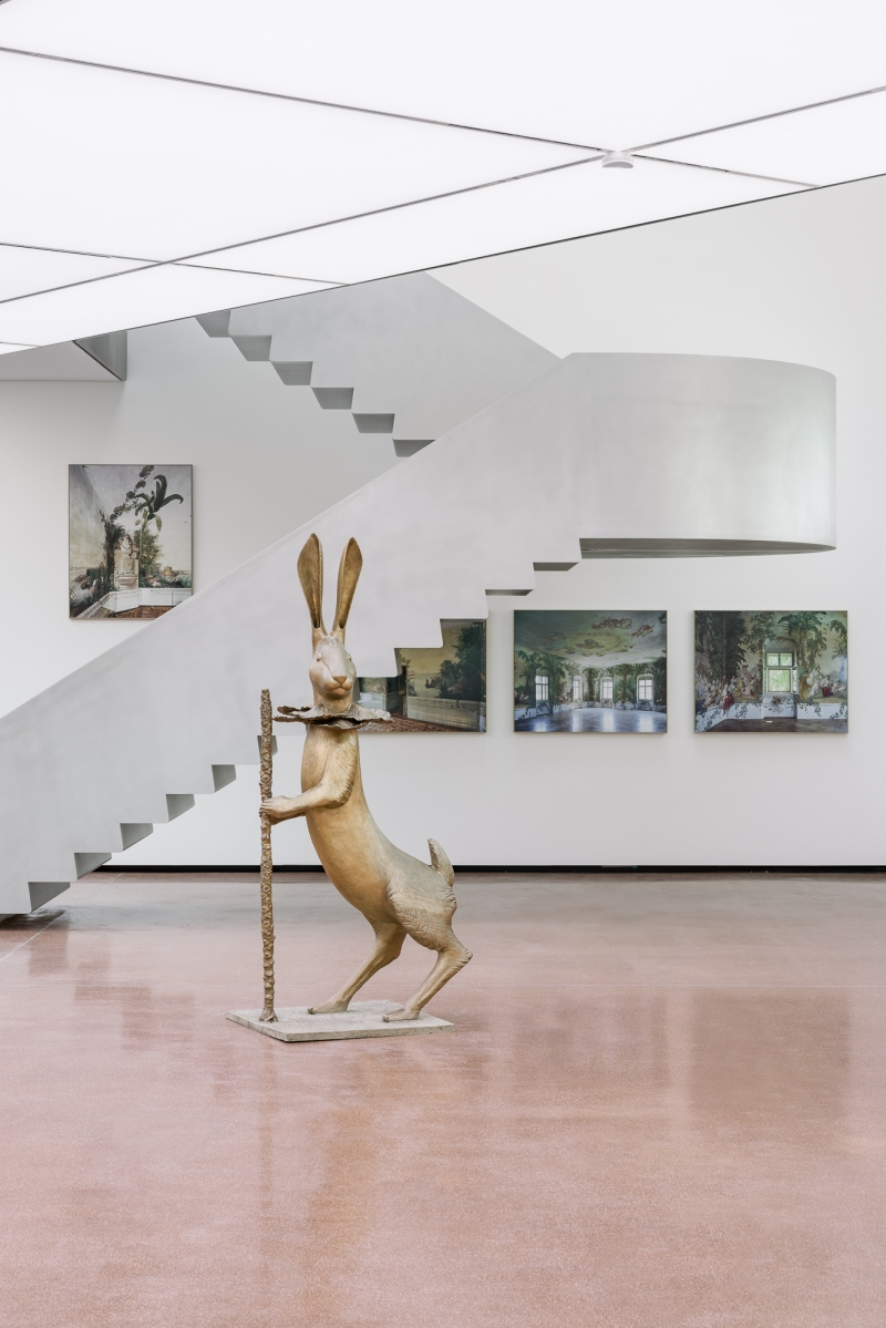 Skulptur eines Hasen mit Stock vor einer geschwungenen Treppe und mehreren Gemälden an der Wand.
