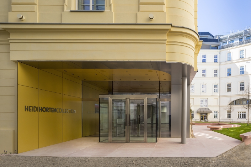 Eingang zur Heidi Horten Collection mit gelber Fassade und gläserner Drehtür, umgeben von einem modernen Innenhof.