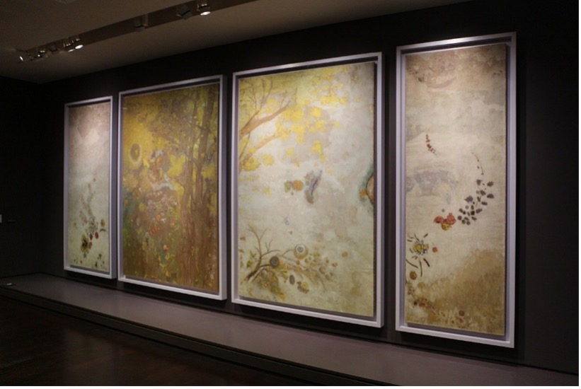 Vier große, gerahmte Gemälde in sanften Farben, hängen an einer dunklen Wand im Musée d'Orsay.