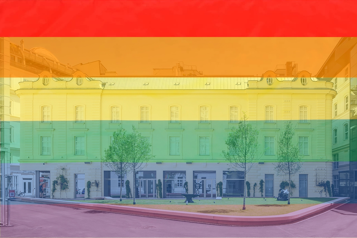 Fassade eines Gebäudes mit Regenbogenfarben im Hintergrund, umgeben von Bäumen und einer kleinen Grünfläche.