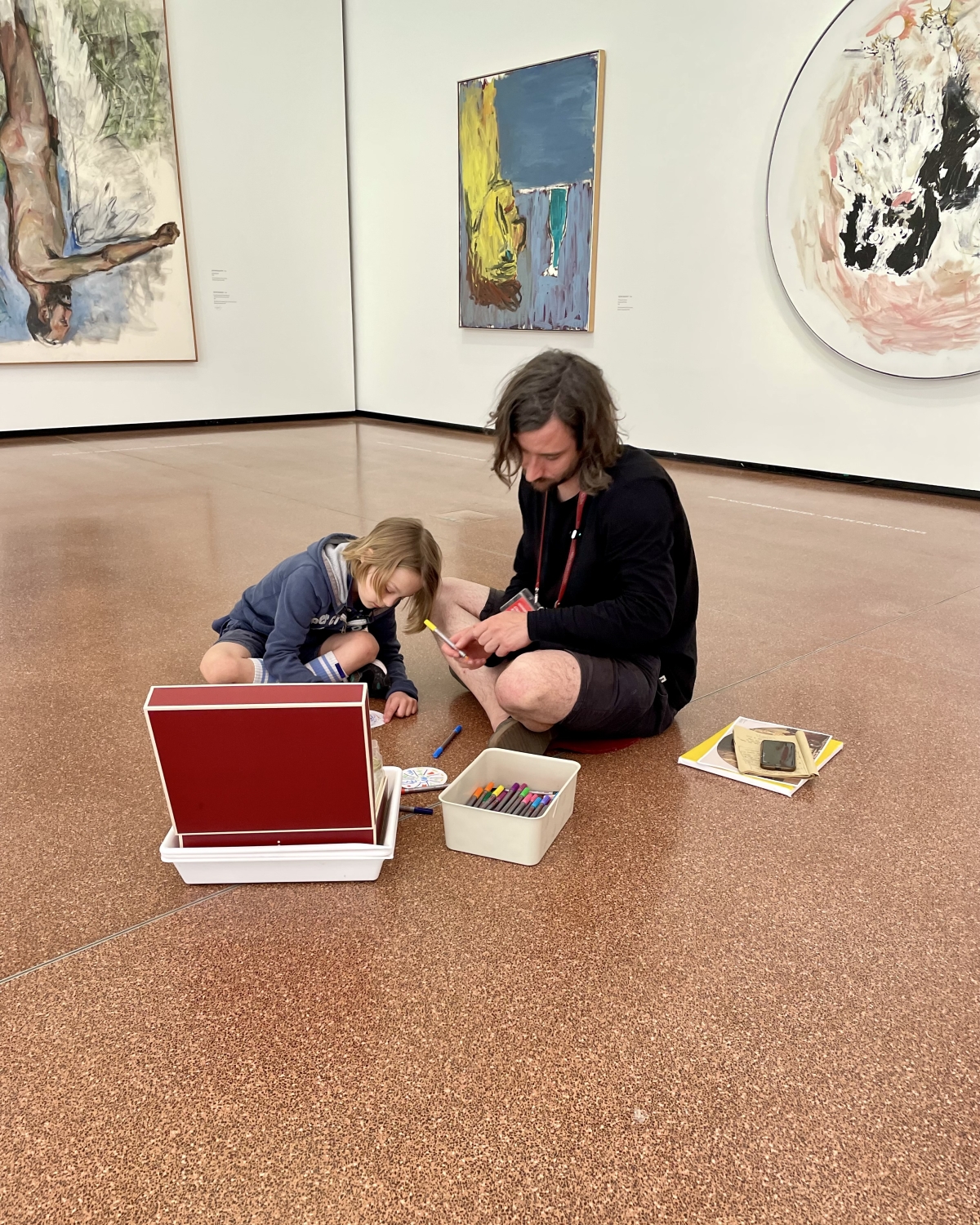 Ein Erwachsener und ein Kind sitzen auf dem Boden eines Kunstmuseums, umgeben von Gemälden, und zeichnen.