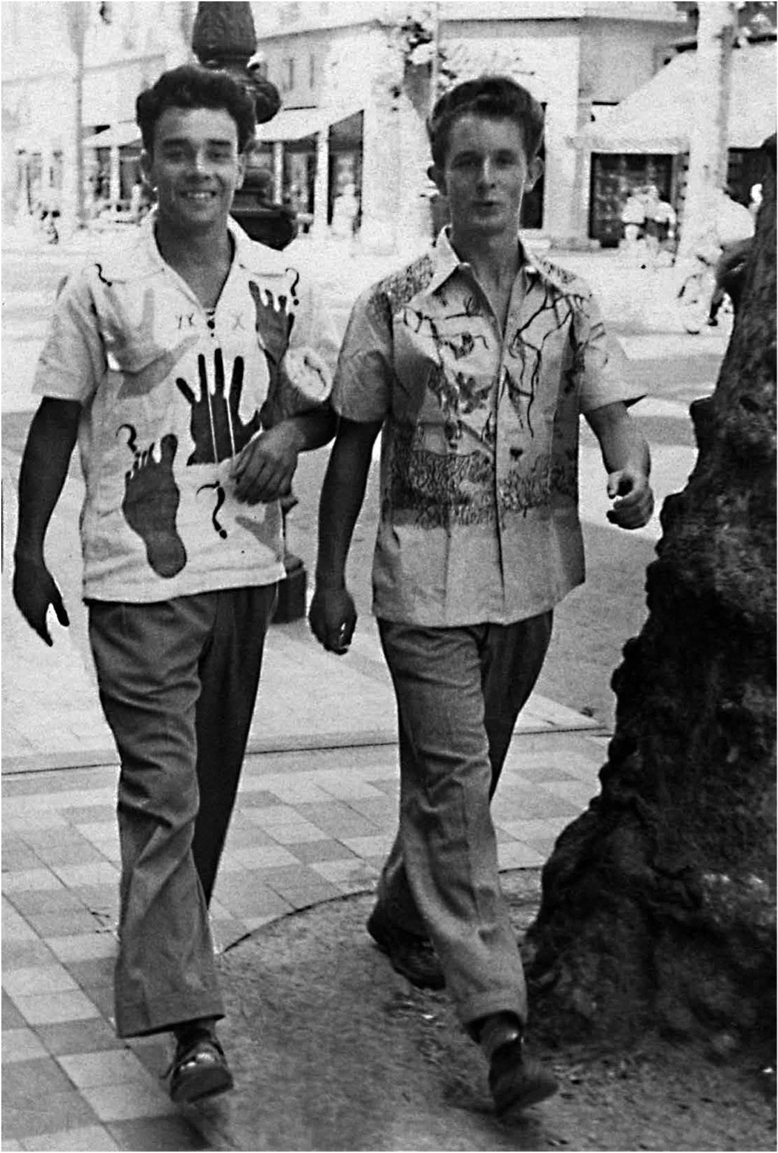 Zwei junge Männer in bunten Hemden gehen lächelnd auf einer Straße entlang, umgeben von Geschäften und Bäumen.