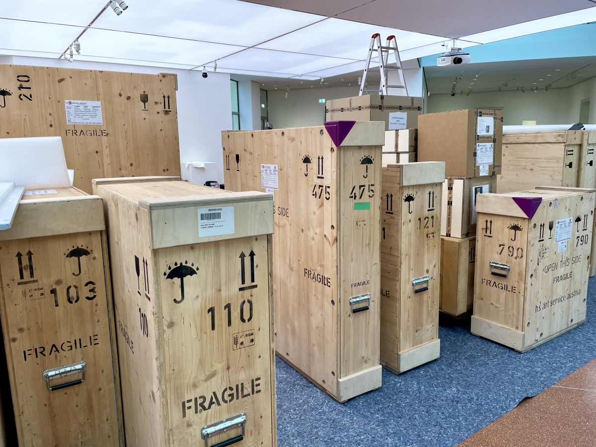 Holzkisten mit Aufschriften und Fragile-Symbolen stehen in einem hellen Raum, im Hintergrund eine Leiter.