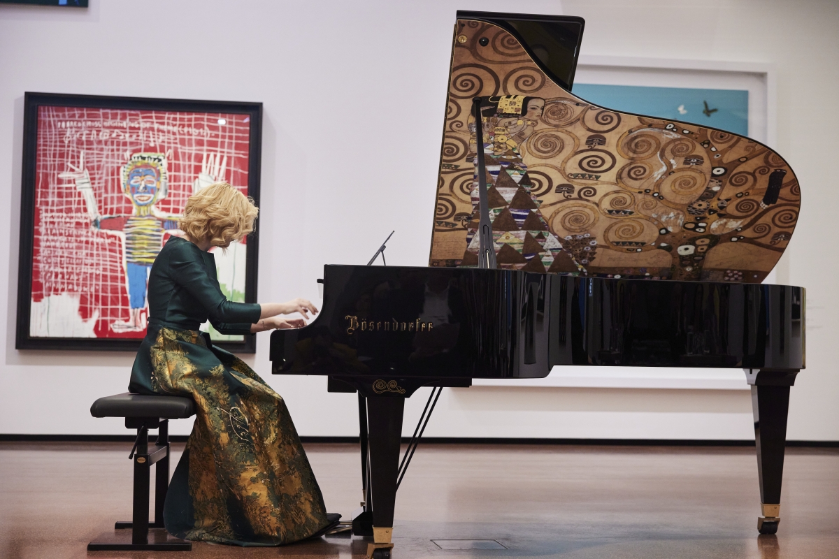 Frau mit lockigem Haar spielt an einem kunstvoll verzierten Klavier in einer Galerie, im Hintergrund moderne Kunstwerke.