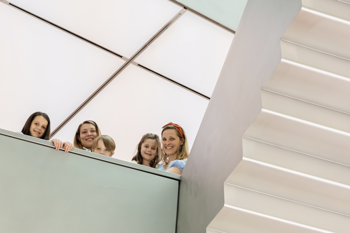 Auf dem Foto sieht man fünf Personen, darunter drei Kinder und zwei Erwachsene. Sie stehen auf einer Ausstellungsebene und schauen freundlich in die Kamera. Alle lächeln.
Rechts im Bild sieht man eine moderne, helle Treppe. Hinter den Personen sieht man die weiße Lichterdecke.