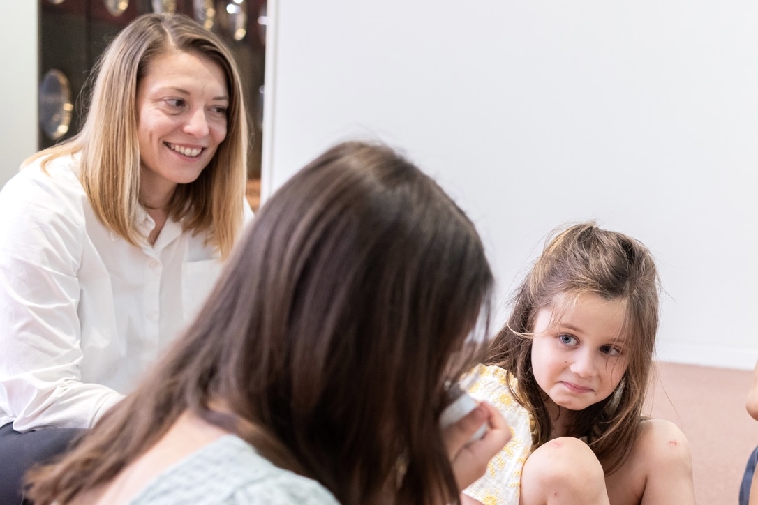 Eine Frau lächelt einer sitzenden, kleinen Mädchen zu, während ein weiteres Mädchen aufmerksam zuhört.