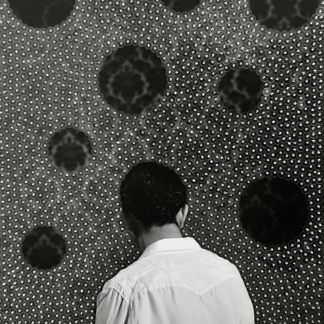 Mann mit schwarzem Haar steht vor einer strukturierten Wand mit runden, dunklen Öffnungen und glänzenden Perlen.