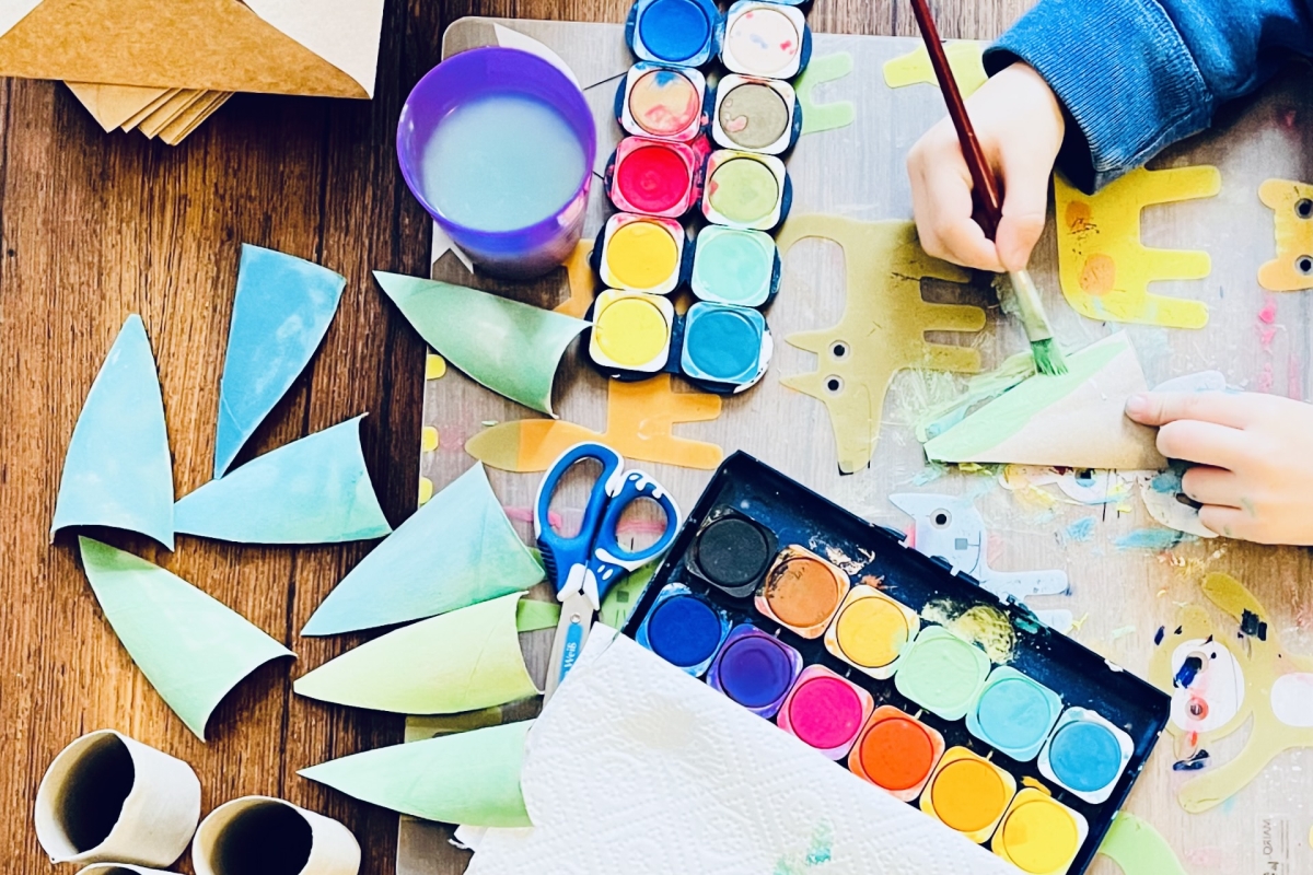 Bunte Bastelmaterialien, Pinsel, Wasserfarben und Papierformen auf einem Tisch, ein Kind malt mit einer Pinsel.