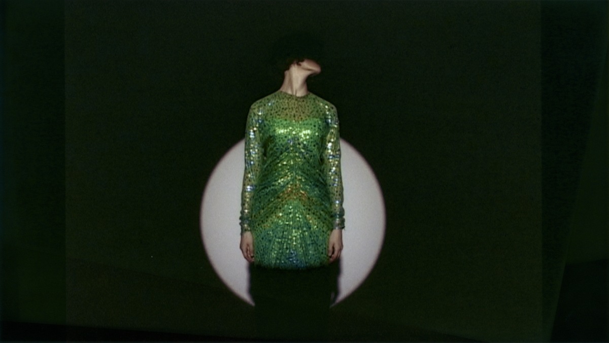 Auf dem Bild sieht man eine Person in einem grünen Kleid.
Das Kleid ist glänzend und mit vielen Pailletten oder Glitzersteinen verziert.
Die Person steht vor einem dunklen Hintergrund,
in dem ein runder, heller Lichtkreis, wahrscheinlich von einem Scheinwerfer, zu sehen ist
Der Kopf der Person ist nach links geneigt,
sodass man das Gesicht nicht sehen kann.
Das macht das Bild geheimnisvoll und spannend.