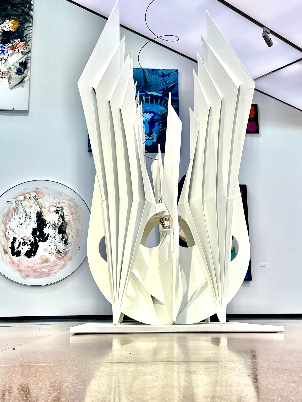 Skulptur mit weißen, spitzen Elementen in einem modernen Ausstellungsraum, umgeben von bunten Gemälden.