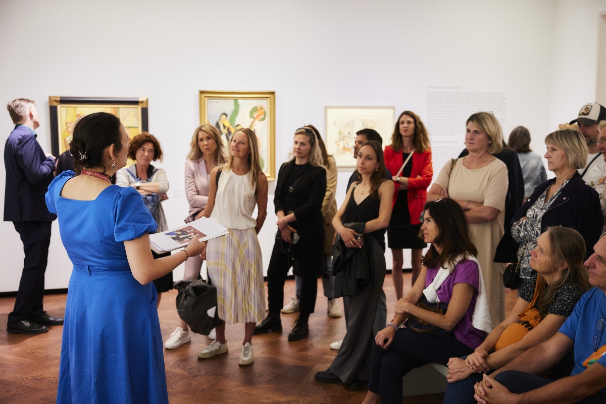 Eine Gruppe von Menschen hört einer Präsentation in einer Kunstgalerie zu, umgeben von Bildern an der Wand.