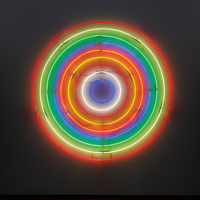 Das Bild zeigt ein leuchtendes Kunstwerk von John M Armleder aus Neonröhren. Mehrere bunte Ringe leuchten in verschiedenen Farben – von innen nach außen: Blau, Weiß, Rot, Orange, Gelb, Grün und Rot. Die Ringe liegen genau übereinander und bilden zusammen eine Zielscheibe oder ein rundes Farbziel. Die Farben leuchten hell vor einem dunklen Hintergrund. Das Licht wirft einen sanften Schein auf die Wand. Das Kunstwerk hängt in einem Ausstellungsraum mit dunklem Holzfußboden.