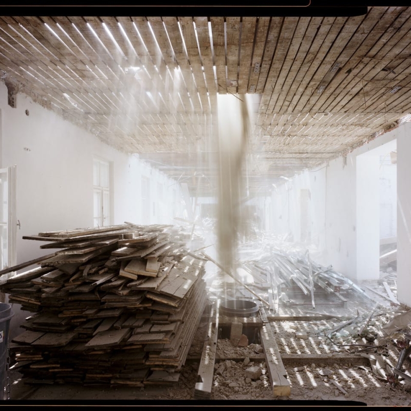 Renovation site with stacked wooden planks, beams of light filtering through a damaged ceiling, and debris scattered on the floor.