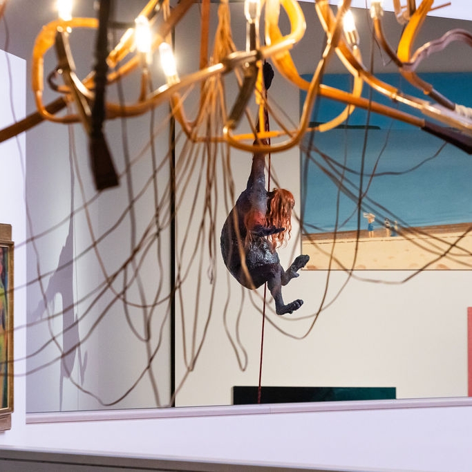 Eine hängende Figur mit langen Haaren schwingt an einem Draht, umgeben von Kunstwerken und einer auffälligen Deckenleuchte.