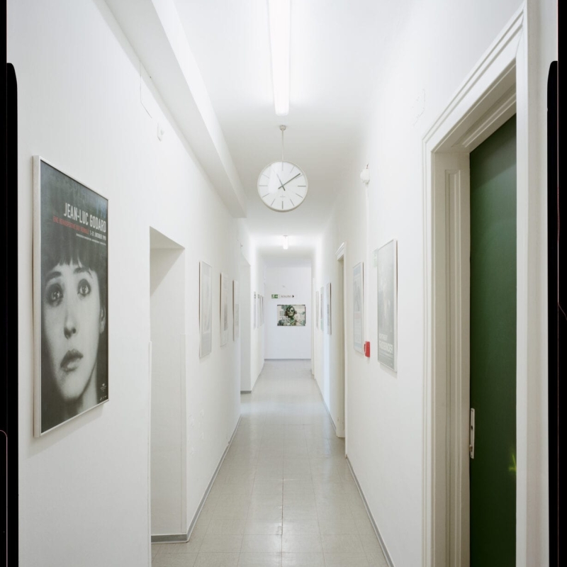 A bright, white hallway lined with framed artworks, featuring a clock and doors on either side.