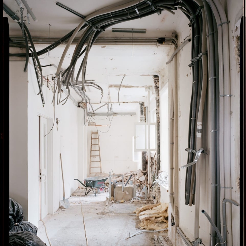 Renovation scene with exposed walls, electrical pipes, a ladder, and construction debris scattered on the floor.