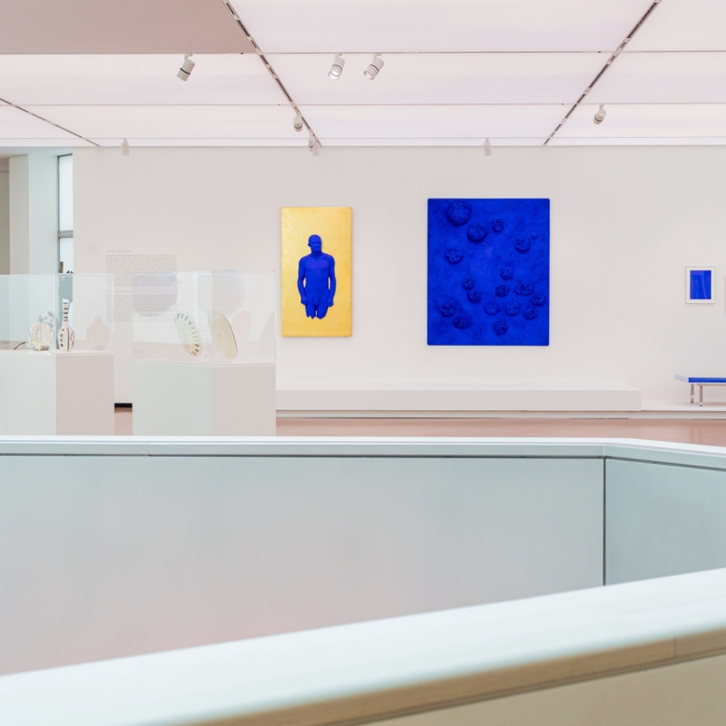 Moderne Galerie mit blauen und goldenen Kunstwerken, Vitrinen mit Skulpturen und minimalistischer Sitzgelegenheit.