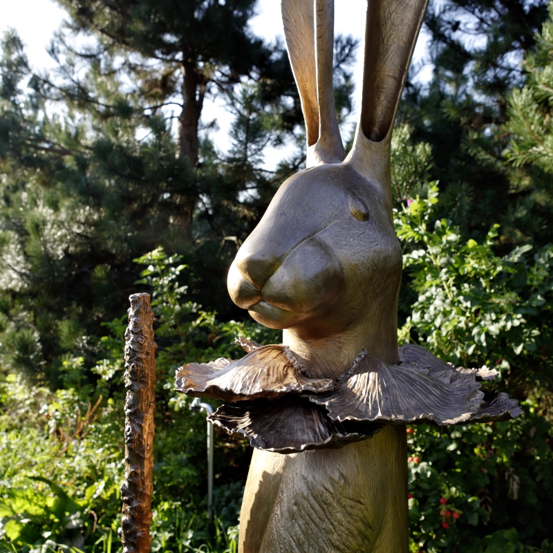 Bronze sculpture of a rabbit with large ears, wearing a ruffled collar, holding a stick, surrounded by greenery.
