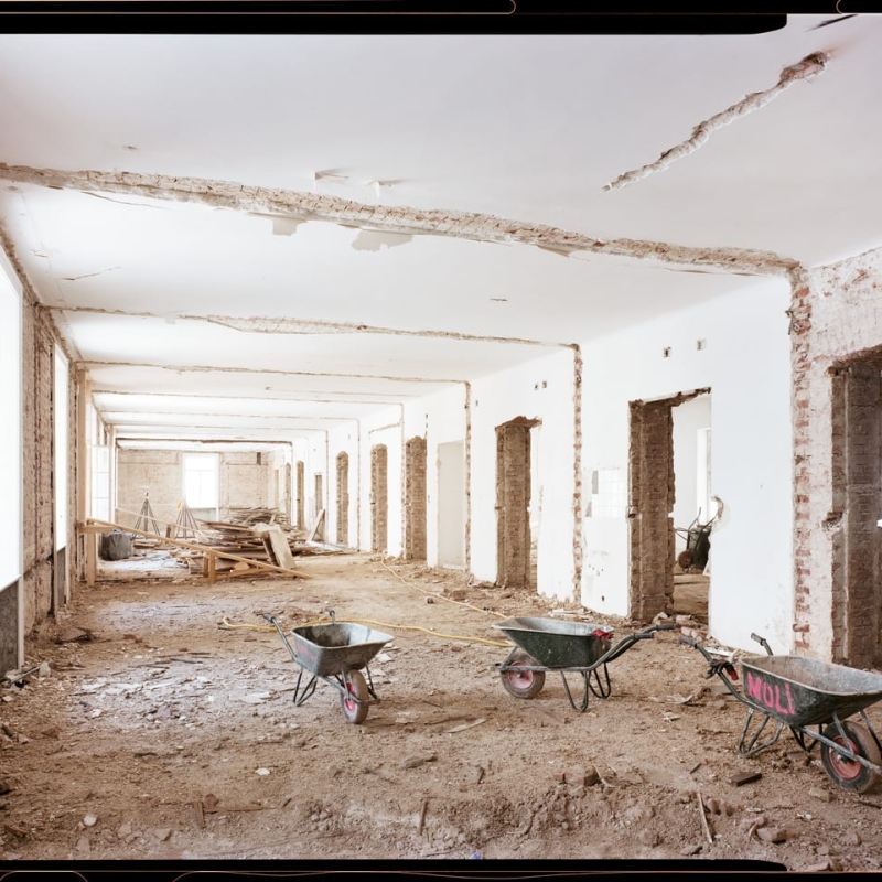 Renovation site with exposed brick walls, three wheelbarrows on dirt floor, and large windows letting in natural light.