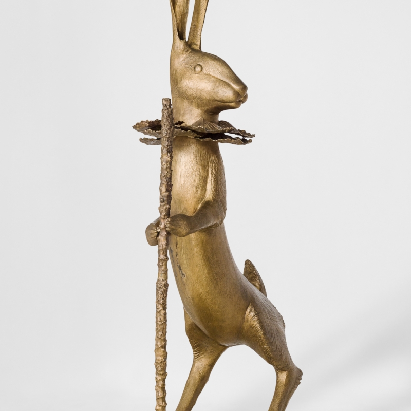 Goldene Hase-Skulptur mit aufrechtem Körper, einem Stock in der Hand und einem Blütenkranz um den Hals.