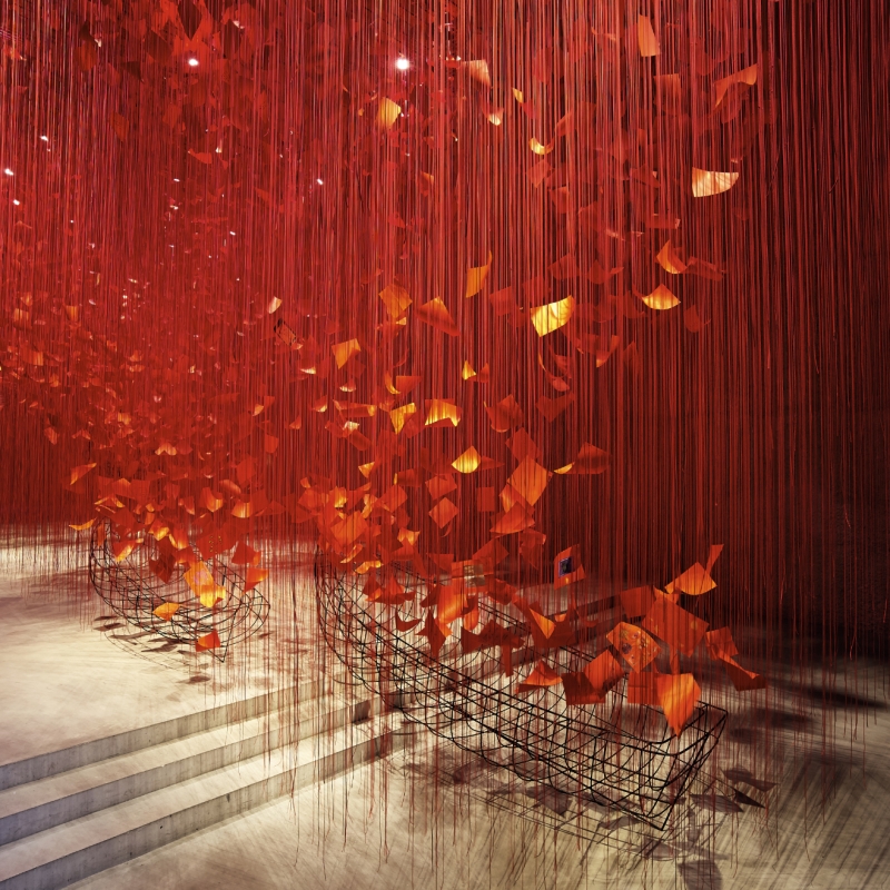 Rote Fäden hängen von der Decke, umgeben von orangefarbenen Blättern und einem Drahtgitter im Hintergrund.
