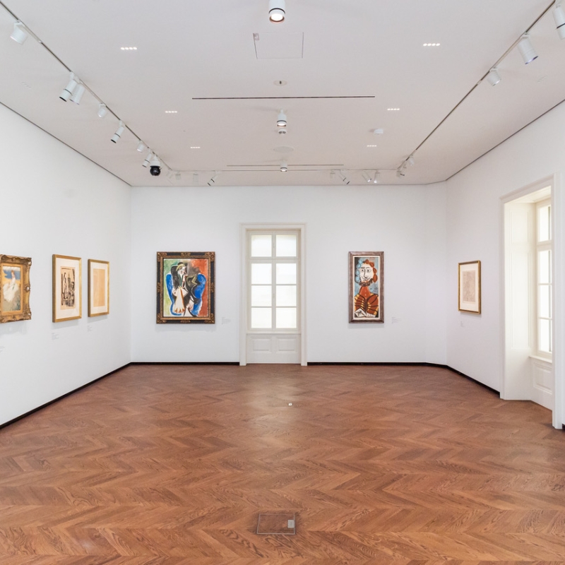 Ausstellungsraum mit mehreren Gemälden an weißen Wänden und einem Fenster mit Holzrahmen. Holzfußboden sichtbar.