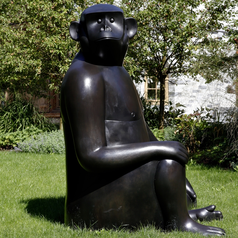 Large bronze sculpture of a seated monkey with a smooth surface, surrounded by green grass and trees.