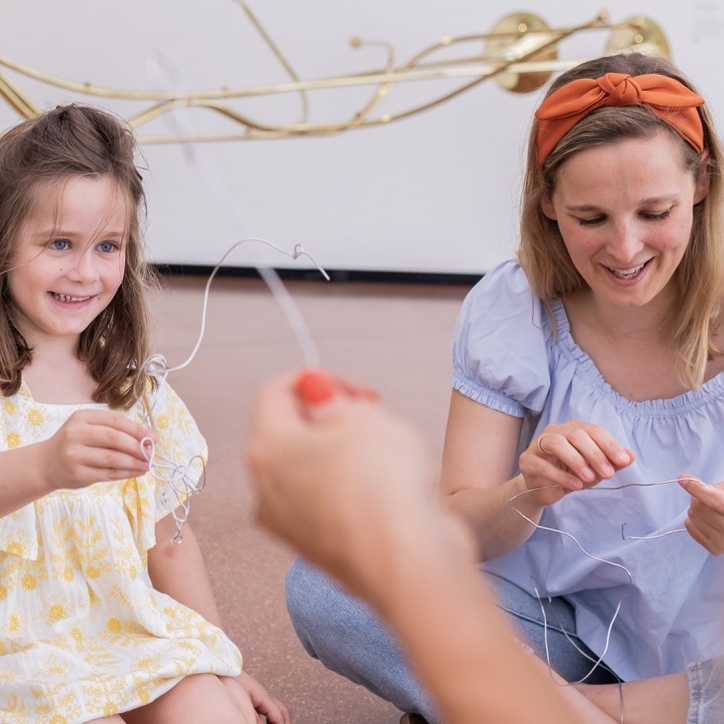 The picture shows a girl and a young woman doing handicrafts together.

Both are sitting on the floor and holding thin wires in their hands.
They are laughing and clearly having fun.
A metal work of art can be seen in the background.

The photo shows a creative hands-on activity.
The atmosphere is cheerful and relaxed.