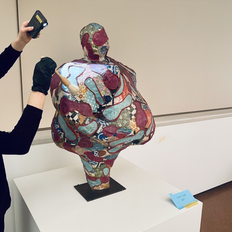 Skulptur eines menschlichen Torso mit bunten, gemusterten Oberflächen, während eine Person sie mit einer Taschenlampe inspiziert.